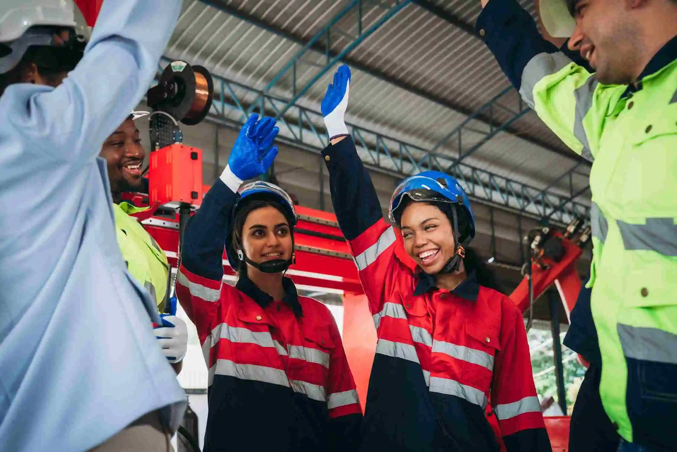 Group of industrial workers wearing safety gear and high-visibility clothing celebrate with high-fives during a team huddle in a manufacturing facility. The joyful atmosphere demonstrates how to keep safety meetings fun while promoting teamwork, engagement, and a strong safety culture on the job site.
