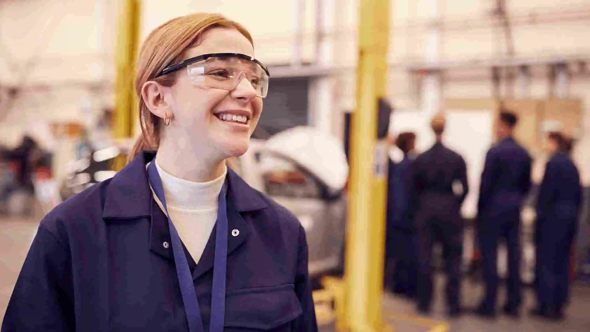 Smiling female worker wearing safety glasses in an industrial training facility, representing simpler safety processes that make workplace practices more effective and easier to follow.